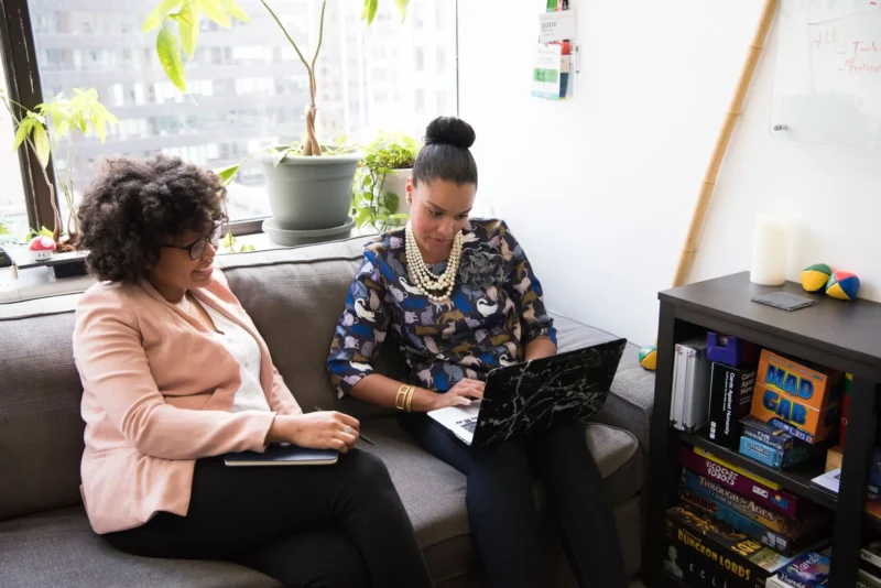 Two women sitting on a couch. One of them using a laptop.